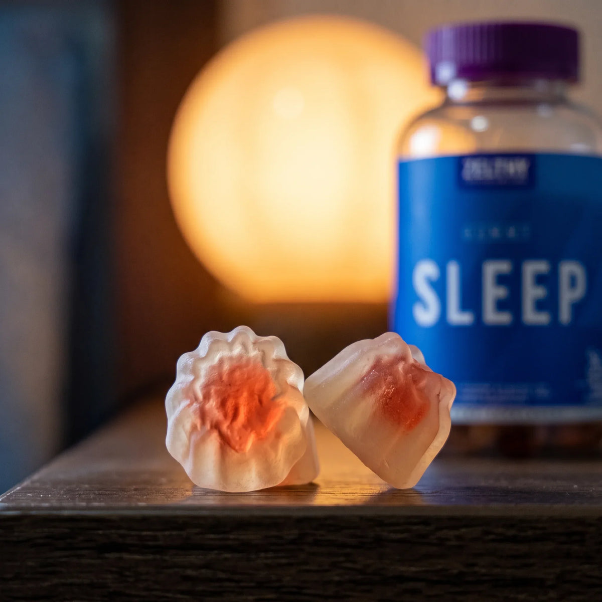 Duas gomas Sleep Zelthy em primeiro plano, com o núcleo softgel visível e o frasco desfocado atrás