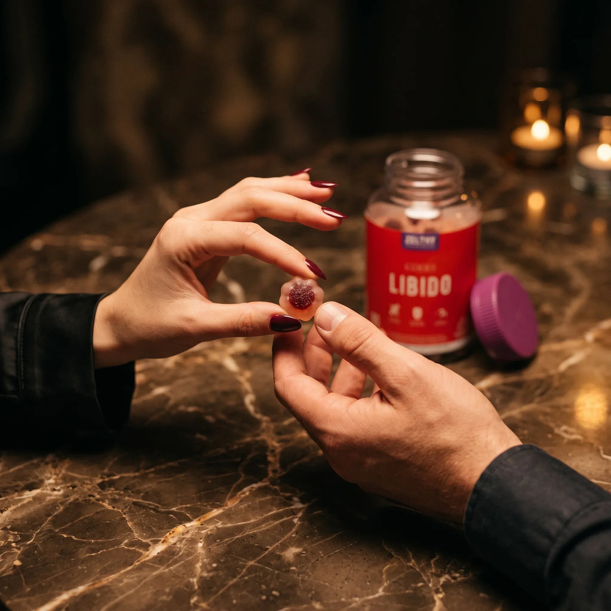 Duas mãos a passar uma goma Libido Zelthy, com o frasco e velas em fundo